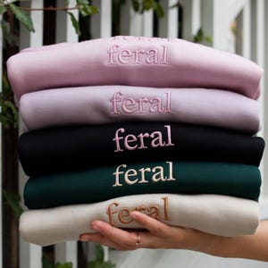 May include: A stack of five sweatshirts in different colors, each with the word "feral" embroidered on the front in a different color.