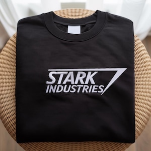 May include: A black crew neck sweatshirt with the white text "STARK INDUSTRIES" and a white angular design. The sweatshirt is folded neatly on a round, woven wicker surface.