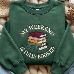 May include: A dark green sweatshirt featuring the embroidered text "MY WEEKEND IS FULLY BOOKED" above a stack of books in various colours. The sweatshirt has a crew neck and long sleeves. The background includes a cream knitted blanket and white trainers.
