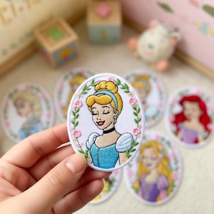 May include: Embroidered oval patches featuring Disney princesses. The patches depict Cinderella, Elsa, Ariel, and Rapunzel, each surrounded by a floral border. Cinderella is in a blue dress, and the patches are on a white background.