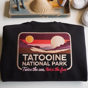 Op de afbeelding: Zwart sweatshirt met een geborduurde patch met een woestijnlandschap met twee zonnen en de tekst "TATOOINE NATIONAL PARK". De tekst "Twice the sun, twice the fun" is ook opgenomen. Andere items in de afbeelding zijn een speelgoedrobot, een zonnebril en witte sneakers.