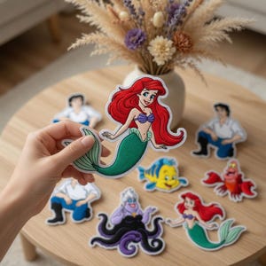 May include: Embroidered patches featuring characters from a popular animated film. The patches include a mermaid with red hair, a crab, a fish, and other characters. The patches are colorful and detailed, with a white border.
