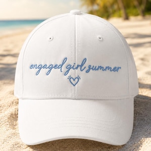 Puede incluir: Una gorra de béisbol blanca con el texto bordado "engaged girl summer" en letra cursiva azul claro, situado sobre el contorno de un pequeño corazón azul. La gorra tiene una corona estructurada, una visera curva y ojales de ventilación.