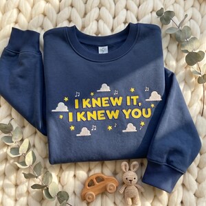 May include: A navy blue crewneck sweatshirt featuring yellow embroidered text that reads "I KNEW IT, I KNEW YOU". The design includes white embroidered clouds, yellow stars, and musical notes scattered around the text on the front of the garment.