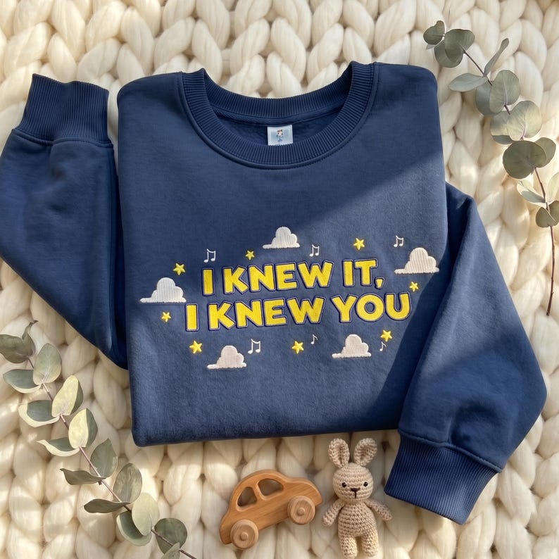 May include: A navy blue crewneck sweatshirt featuring yellow embroidered text that reads "I KNEW IT, I KNEW YOU". The design includes white embroidered clouds, yellow stars, and musical notes scattered around the text on the front of the garment.