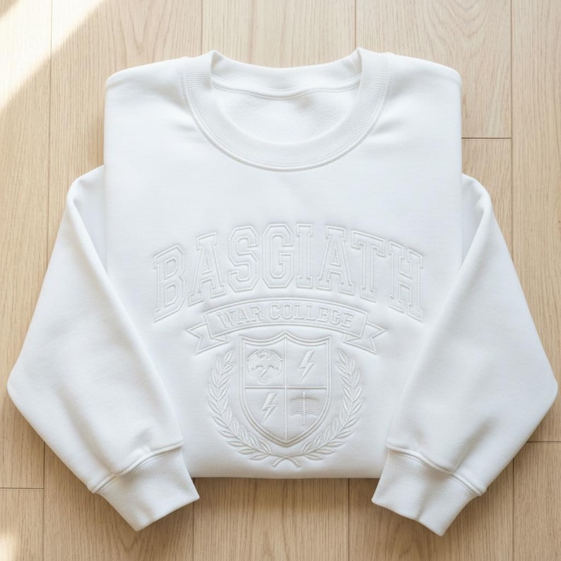 Może przedstawiać: Biała bluza z okrągłym dekoltem z wytłoczonym napisem "BASGIATH WAR COLLEGE" i herbem. Bluza jest złożona na jasnej drewnianej powierzchni. Projekt zawiera tarczę, błyskawice i wieniec laurowy.