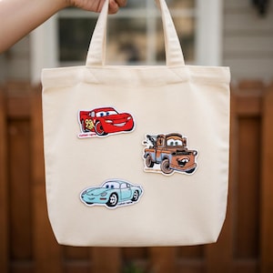 May include: A beige canvas tote bag featuring three colorful car patches: a red race car with the number 95, a brown tow truck, and a light blue sports car. The bag has two handles.