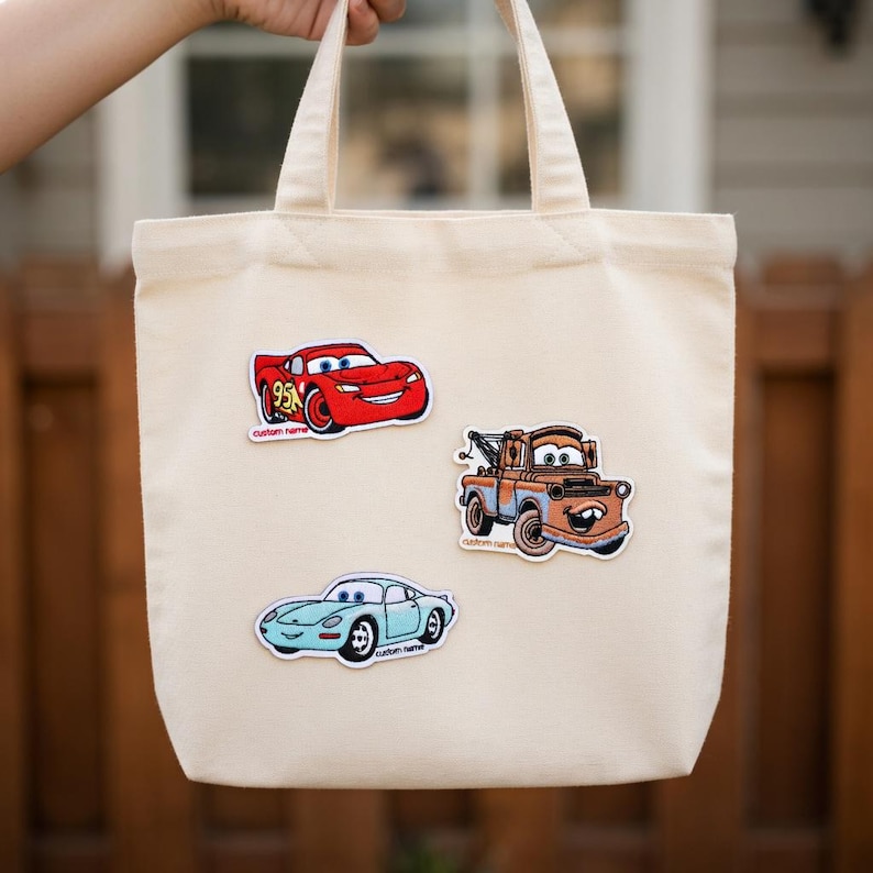 May include: A beige canvas tote bag featuring three colorful car patches: a red race car with the number 95, a brown tow truck, and a light blue sports car. The bag has two handles.