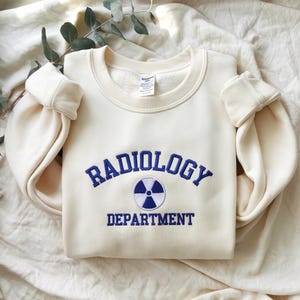 Könnte beinhalten: Cremefarbenes Sweatshirt mit dem blau gestickten Schriftzug "RADIOLOGY DEPARTMENT". Ein blau-weißes Strahlungssymbol befindet sich zwischen den Wörtern. Das Sweatshirt hat einen Rundhalsausschnitt und gerippte Bündchen.