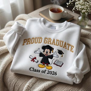May include: White sweatshirt with "PROUD GRADUATE" in gold lettering and a cartoon Mickey Mouse in a graduation gown. The sweatshirt also features "Class of 2026" in black text. A graduation-themed gift.
