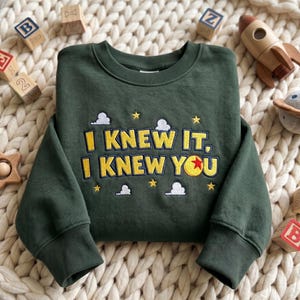 May include: A dark green long-sleeve sweatshirt featuring embroidered yellow text that reads I KNEW IT, I KNEW YOU. The design includes small white clouds and yellow stars, with a red, yellow, and blue ball replacing the letter O.