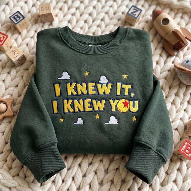 May include: A dark green long-sleeve sweatshirt featuring embroidered yellow text that reads I KNEW IT, I KNEW YOU. The design includes small white clouds and yellow stars, with a red, yellow, and blue ball replacing the letter O.