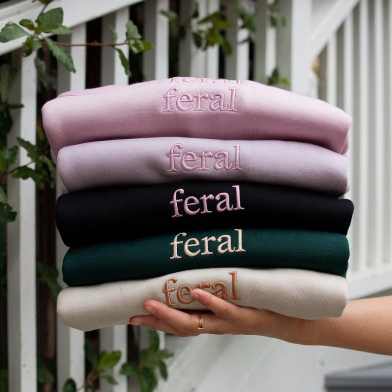 May include: A stack of five sweatshirts in different colors, each with the word "feral" embroidered on the front in a different color.