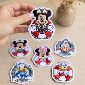 May include: A collection of embroidered patches featuring cartoon characters dressed as sailors, including Mickey and Minnie Mouse, Donald Duck, and Goofy. Each patch is set within a red and white life preserver design. The patches are colourful and detailed.