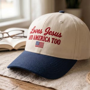 Puede incluir: Una gorra de béisbol de color crema con visera azul marino. La parte frontal presenta un texto bordado en rojo que dice "Loves Jesus AND AMERICA TOO" sobre una pequeña bandera estadounidense rectangular bordada con rayas rojas, blancas y azules.