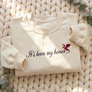Puede incluir: Sudadera color crema con la frase bordada "It's been my honor" en escritura azul marino, acompañada de un emblema de dragón rojo. La sudadera tiene cuello redondo y puños acanalados.