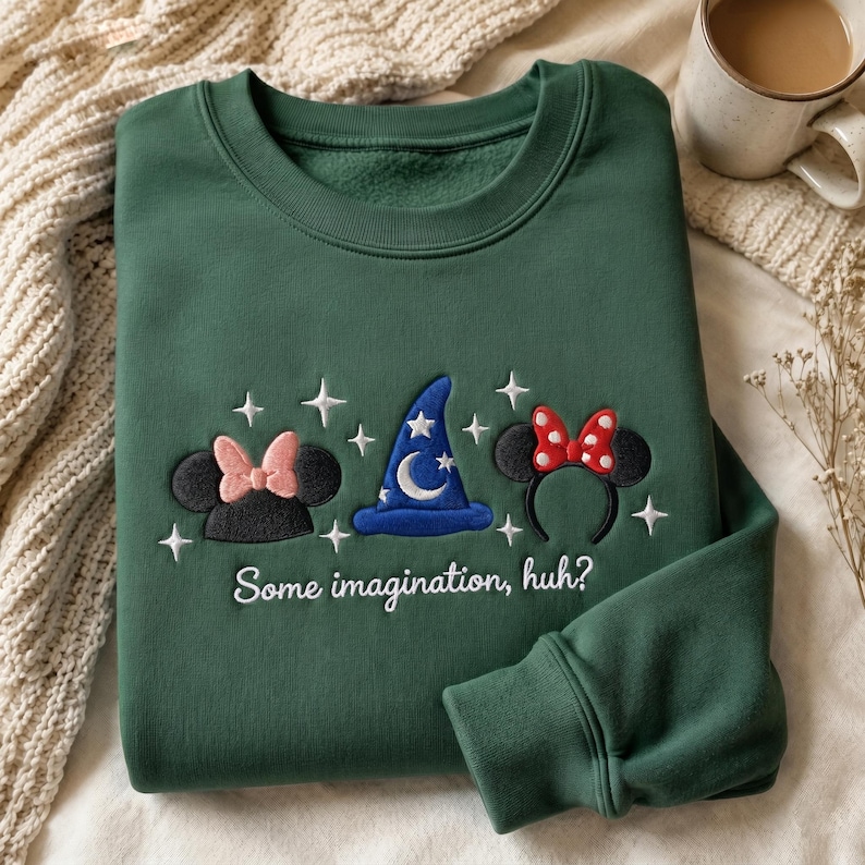 May include: A forest green crewneck sweatshirt featuring embroidered designs of a black mouse ear headband with a pink bow, a blue wizard hat with a white moon and stars, and a black mouse ear headband with a red polka dot bow, with the text "Some imagination, huh?"