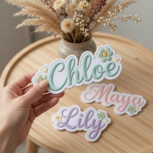 Pode incluir: Patches bordados com nomes em cores pastel. Os nomes "Chloe", "Maya" e "Lily" estão em verde claro, rosa e lavanda. Cada patch apresenta motivos florais e de coração. Os patches estão em uma superfície de madeira, com um vaso de flores secas ao fundo.