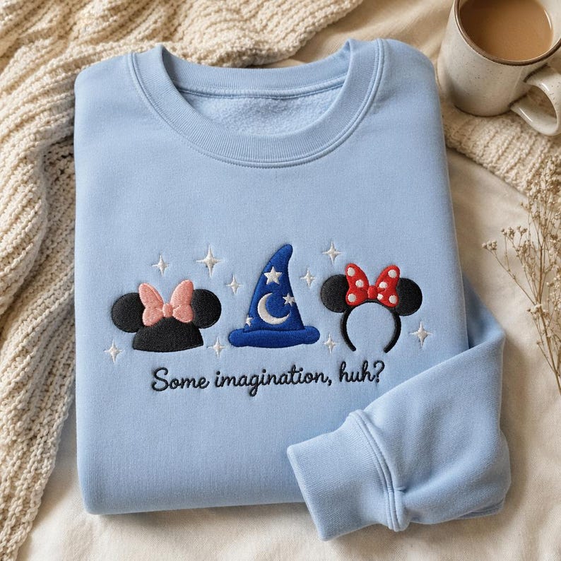 May include: A light blue crewneck sweatshirt featuring embroidered designs of black mouse ears with a pink bow, a blue wizard hat with a white moon and stars, and black mouse ears with a red polka dot bow, with the text "Some imagination, huh?"
