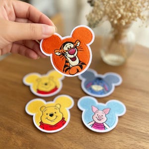 May include: Embroidered patches featuring Disney characters, including Tigger, Winnie the Pooh, Eeyore, and Piglet. The patches have a Mickey Mouse head shape with orange, yellow, blue, and pink colors. The Tigger patch is held up by a hand.