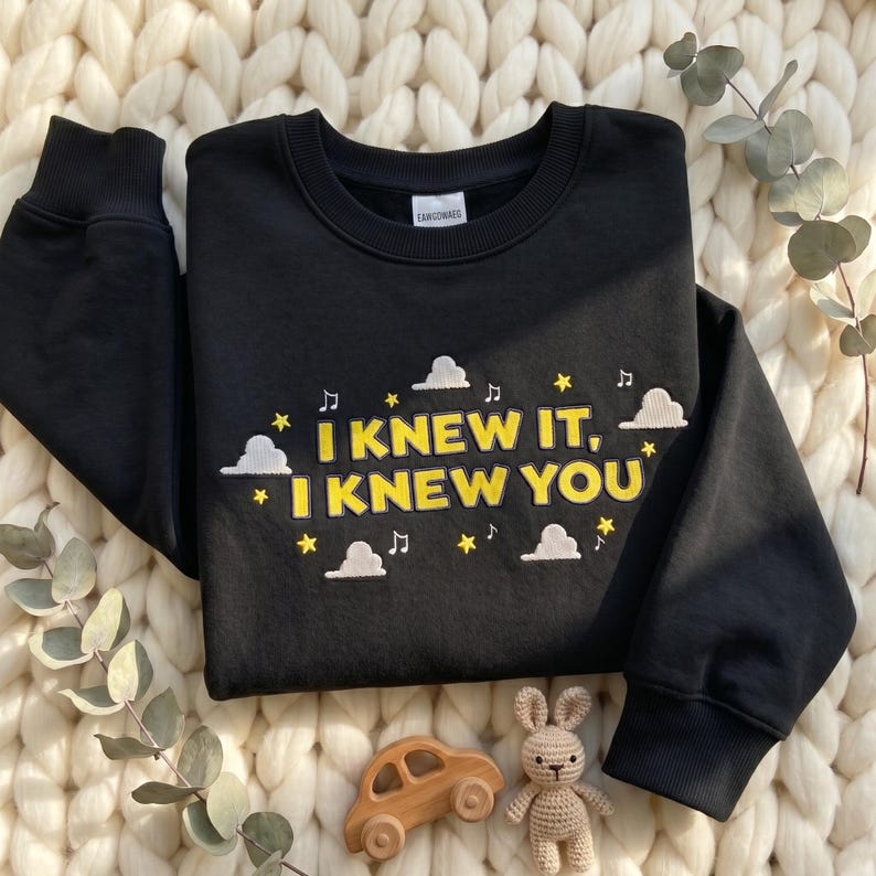 May include: A black long-sleeve sweatshirt featuring yellow embroidered text that reads I KNEW IT, I KNEW YOU. The design includes white embroidered clouds, yellow stars, and musical notes scattered around the text on the front of the garment.