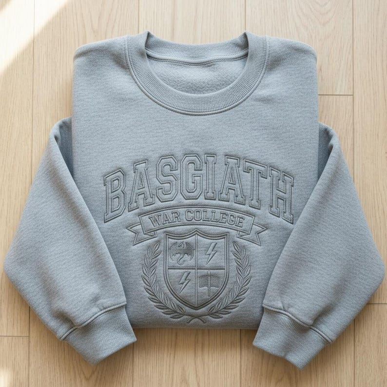 Może przedstawiać: Jasnoszara bluza z napisem "BASGIATH WAR COLLEGE" w wypukłym, teksturowanym wzorze. Poniżej herb z tarczą, piorunami i książką. Bluza jest starannie złożona na drewnianej powierzchni.