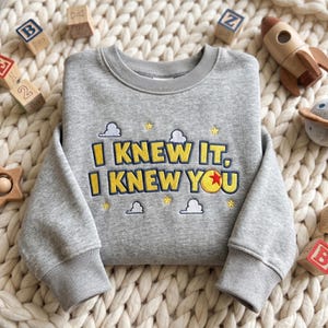 May include: A heather gray crewneck sweatshirt featuring embroidered text that reads I KNEW IT, I KNEW YOU in yellow with dark blue outlines. The design includes small white clouds, yellow stars, and a yellow ball with a red star center.