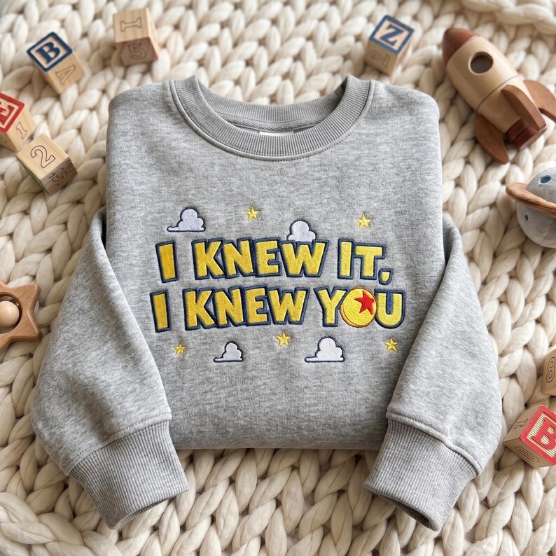 May include: A heather gray crewneck sweatshirt featuring embroidered text that reads I KNEW IT, I KNEW YOU in yellow with dark blue outlines. The design includes small white clouds, yellow stars, and a yellow ball with a red star center.