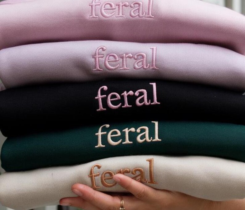 May include: Five sweatshirts stacked on top of each other. The sweatshirts are in different colors: pink, light purple, black, dark green, and beige. Each sweatshirt has the word "feral" embroidered on the front in a different color.