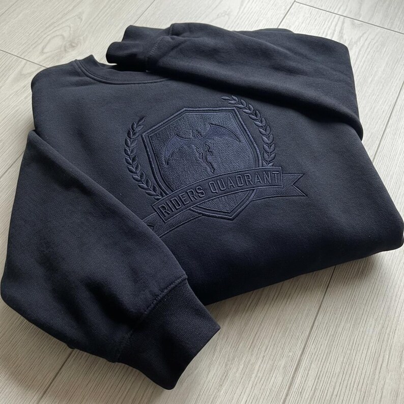 May include: Navy blue sweatshirt with a crew neck. The front features an embroidered emblem with the text "RIDERS QUADRANT" on a banner. The emblem includes a shield, laurel wreath, and a winged figure. The sweatshirt has ribbed cuffs and hem.
