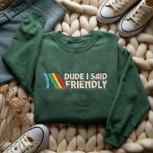 Pode incluir: Uma camisola verde escuro com o texto bordado "DUDE I SAID FRIENDLY" em branco. O design inclui um arco-íris. A camisola é exibida com jeans e tênis de lona.