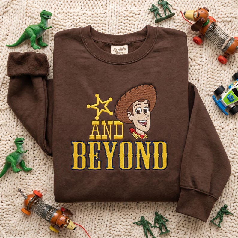 May include: A dark brown crewneck sweatshirt featuring an embroidered graphic of a cowboy character, a yellow star, and the yellow text "AND BEYOND". The interior neck label reads "Andy's Toys". The sweatshirt has long sleeves and ribbed cuffs.