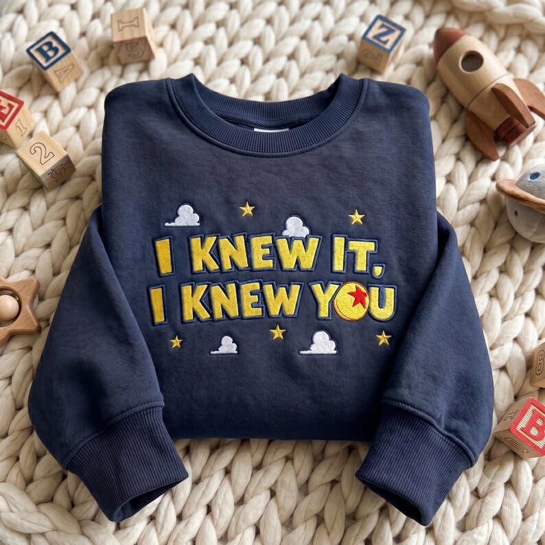 May include: A navy blue long-sleeve sweatshirt featuring yellow embroidered text that reads I KNEW IT, I KNEW YOU. The design includes small white clouds and yellow stars, with a red, yellow, and blue ball replacing the letter O in YOU.