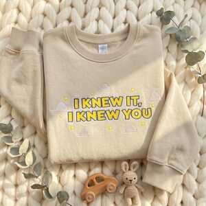May include: A beige long-sleeve sweatshirt featuring embroidered text that reads I KNEW IT, I KNEW YOU in yellow with a dark blue outline. The design includes small embroidered yellow stars, white clouds, and musical notes surrounding the text.