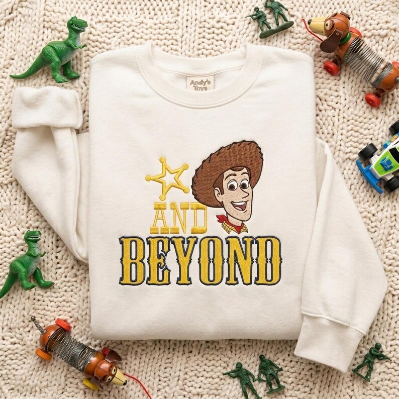 May include: A cream-colored crewneck sweatshirt featuring an embroidered graphic of a cowboy character wearing a brown hat and red neckerchief, accompanied by a yellow star and the yellow embroidered text AND BEYOND in a bold serif font.