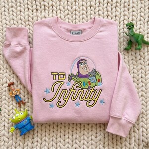 Puede incluir: Una sudadera de manga larga de color rosa claro con un bordado de Buzz Lightyear de Pixar. El dise&ntilde;o incluye el texto "TO Infinity" en letras amarillas, rodeado de peque&ntilde;as estrellas azules bordadas sobre la tela.