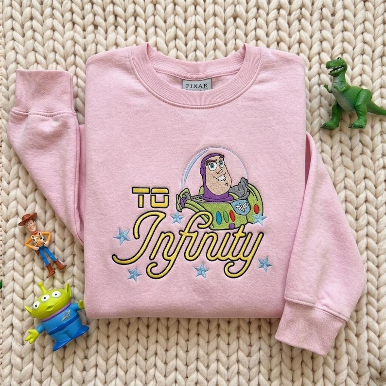 Puede incluir: Una sudadera de manga larga de color rosa claro con un bordado de Buzz Lightyear de Pixar. El dise&ntilde;o incluye el texto "TO Infinity" en letras amarillas, rodeado de peque&ntilde;as estrellas azules bordadas sobre la tela.