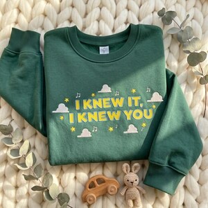 May include: A forest green crewneck sweatshirt featuring embroidered yellow text that reads I KNEW IT, I KNEW YOU. The design includes white clouds, yellow stars, and small musical notes surrounding the text, with ribbed cuffs and a collar.