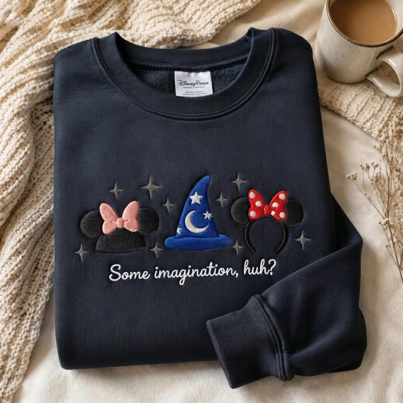 May include: A navy blue crewneck sweatshirt featuring embroidered Disney Parks branding on the inner collar. The front displays a blue wizard hat, two pairs of mouse ears with bows, and the white embroidered text Some imagination, huh?
