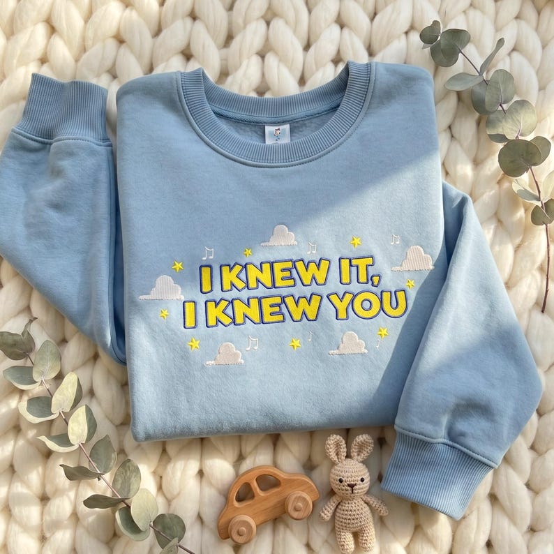 May include: A light blue crewneck sweatshirt featuring embroidered text that reads I KNEW IT, I KNEW YOU in yellow with a dark blue outline. The design includes small white clouds, yellow stars, and musical notes scattered around the lettering.