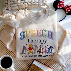 Op de afbeelding: Een crèmekleurige sweater met ronde hals en geborduurde tekst "SPEECH Therapy" in kleurrijke blokletters. Onder de tekst staat een borduurwerk van Winnie de Poeh en vrienden, met de tekst "A LITTLE KINDNESS GOES A LONG WAY" eronder.
