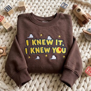 May include: A dark brown long-sleeve sweatshirt featuring embroidered yellow text that reads I KNEW IT, I KNEW YOU. The design includes small white clouds and yellow stars, with a red, yellow, and blue ball replacing the letter O in YOU.