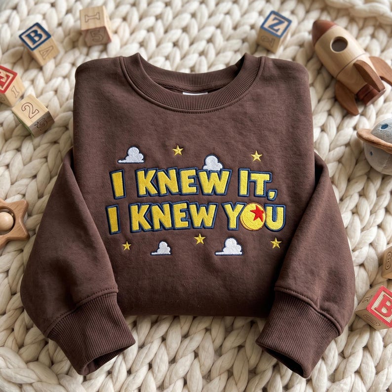 May include: A dark brown long-sleeve sweatshirt featuring embroidered yellow text that reads I KNEW IT, I KNEW YOU. The design includes small white clouds and yellow stars, with a red, yellow, and blue ball replacing the letter O in YOU.