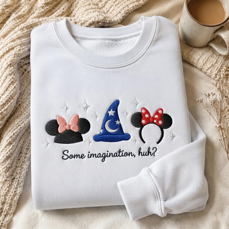 May include: A white long-sleeve sweatshirt featuring embroidered designs of black mouse ears with a pink bow, a blue wizard hat with a white moon and stars, and black mouse ears with a red polka dot bow, with the text "Some imagination, huh?"