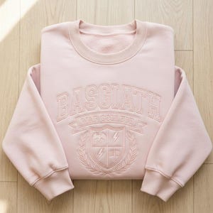 Może przedstawiać: Jasnor&oacute;żowa bluza z haftowanym napisem "BASGIATH WAR COLLEGE" na piersi. Projekt zawiera herb ze smokiem, błyskawicami i tarczą. Bluza jest starannie złożona na drewnianej powierzchni.