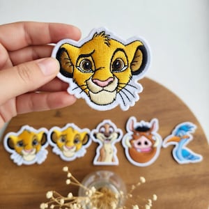Könnte beinhalten: Gestickte Aufnäher mit Charakteren aus Der König der Löwen. Die Aufnäher zeigen Simba, Timon, Pumbaa und Zazu. Die Aufnäher sind gelb, braun, rosa und blau mit weißen Rändern. Die Aufnäher befinden sich auf einer Holzoberfläche.