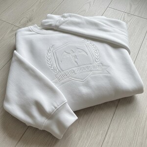 May include: White crewneck sweatshirt with a detailed embroidered emblem. The emblem features a winged figure within a shield, surrounded by a laurel wreath, and the text "RIDERS QUADRANT" on a banner. The sweatshirt is folded on a light wood surface.