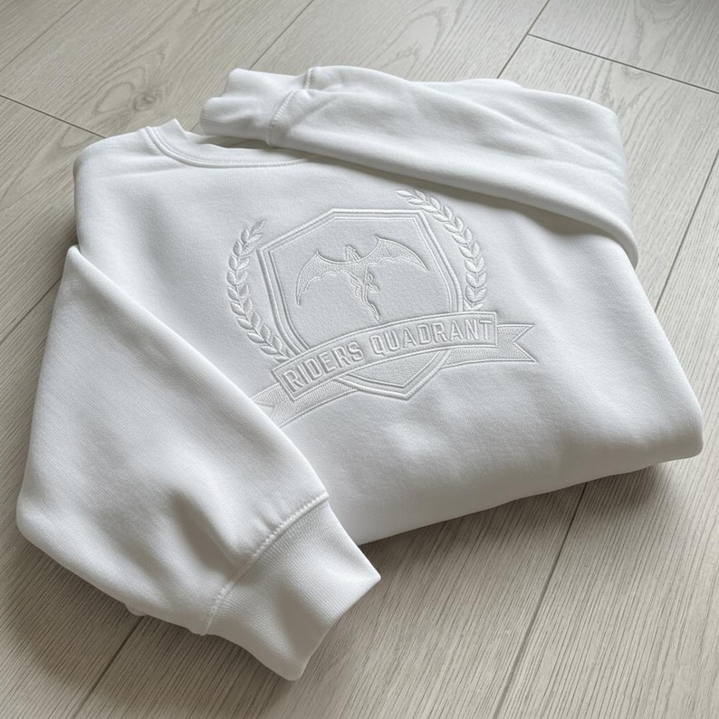 May include: White crewneck sweatshirt with a detailed embroidered emblem. The emblem features a winged figure within a shield, surrounded by a laurel wreath, and the text "RIDERS QUADRANT" on a banner. The sweatshirt is folded on a light wood surface.