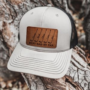 Può includere: Cappellino da camionista grigio chiaro e nero con toppa in pelle. La toppa presenta mazze da golf e il testo "Emma, Noah, Olivia, Liaia, Ava" sopra "BEST DAD BY PAR". Il cappellino ha una visiera curva e un retro in rete.