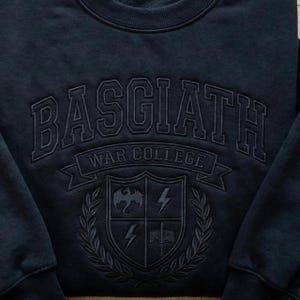 Może przedstawiać: Granatowa bluza z napisem "BASGIATH WAR COLLEGE" w łuku. Poniżej herb z smokiem, piorunami i otwartą książką. Projekt jest w tonacji ton w ton.